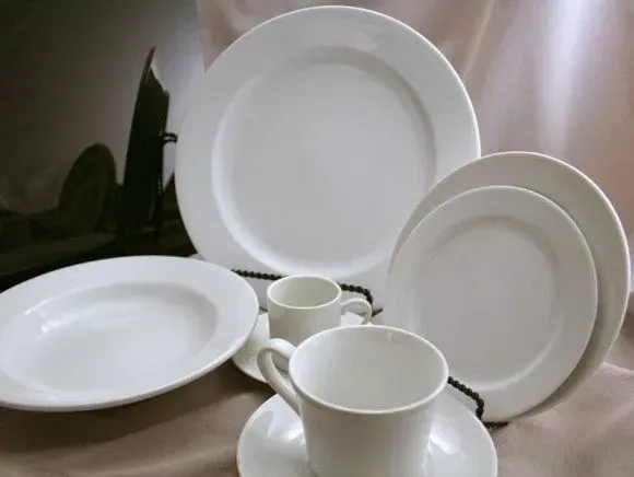 A collection of white ceramic dinnerware, including various plates, saucers, and cups, arranged on a beige surface.