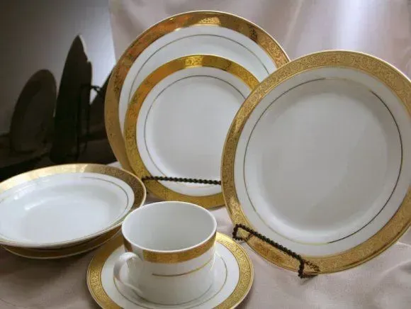 A place setting of white porcelain dishes with wide, textured gold rims and thin gold accent rings on a neutral surface.