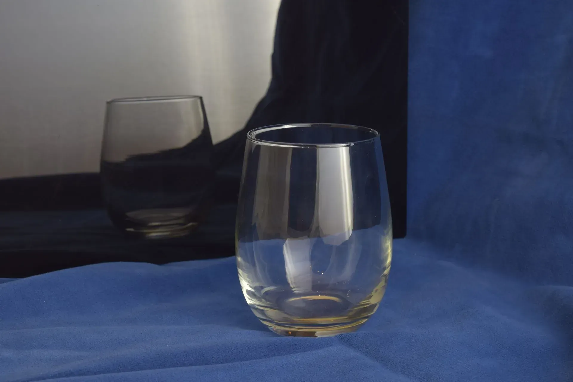 Two clear glasses sit on a blue cloth, with one glass in the foreground and a darker reflection in the background.
