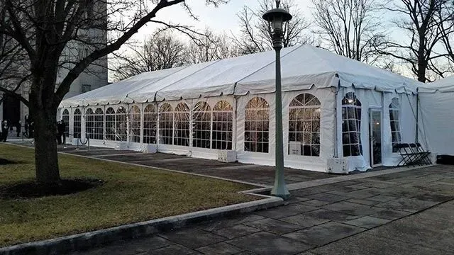 A long, white temporary tent with arched windows set up on a paved patio in a park near bare trees.