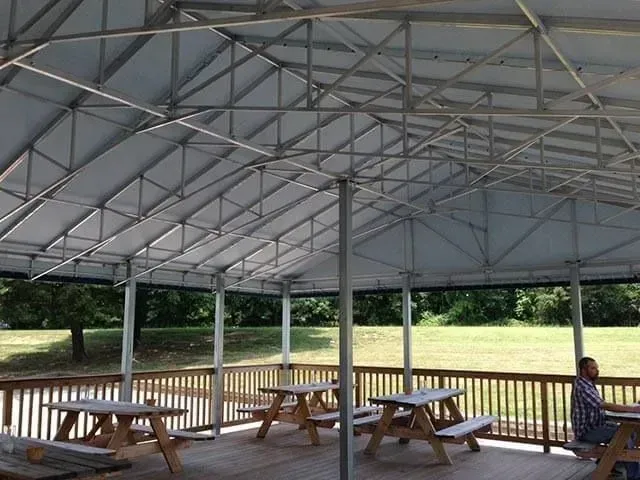 A metal-framed pavilion with a white canopy roof featuring wooden picnic tables on a deck overlooking a grassy field.