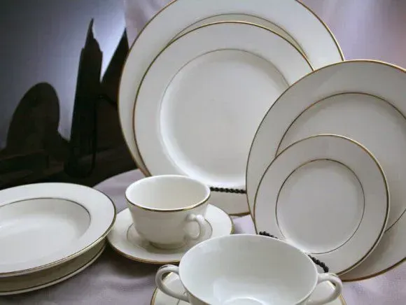 A set of elegant white ceramic dishes with gold rims, including dinner plates, bowls, and teacups, arranged on a cloth.