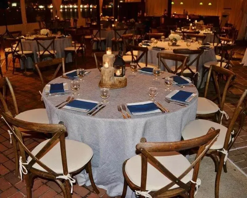 A table with light gray tablecloths and dark blue napkins, set for a formal outdoor evening event with rustic wood chairs.