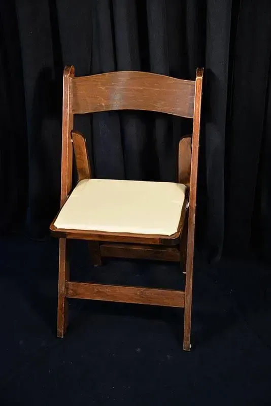 A wooden folding chair with a beige padded seat, centered against a black curtain background.