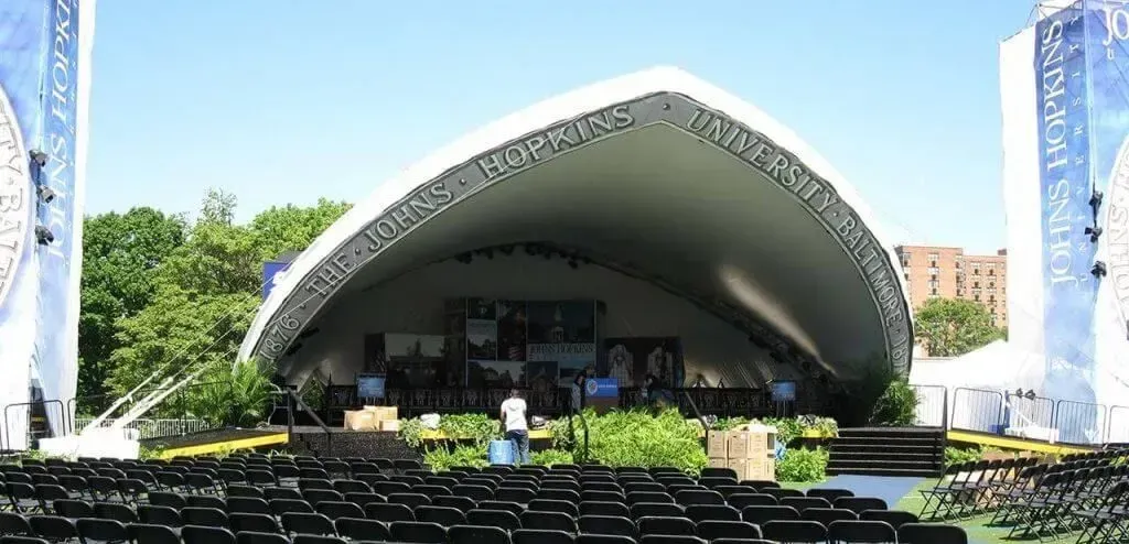 A large outdoor stage with a white arched roof, surrounded by trees and empty rows of black chairs under a blue sky.