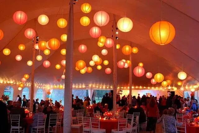 Indoor event tent with many glowing orange, white, and yellow paper lanterns hanging from the ceiling above seated guests.