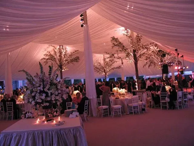 A formal event held in a large, draped tent with white table settings, tall floral centerpieces, and decorative trees.