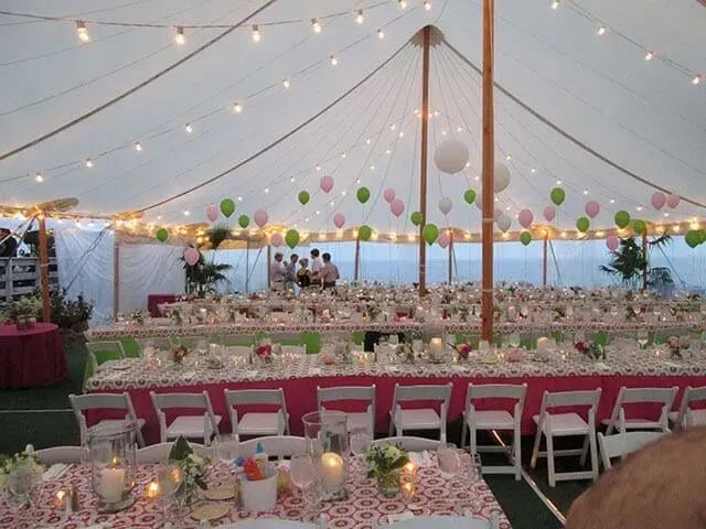 A tented outdoor event with banquet tables, white folding chairs, and hanging green and pink balloons with string lights.