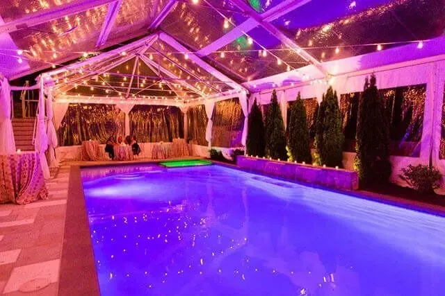 An outdoor swimming pool glows purple at night inside a clear-topped tent illuminated by string lights.