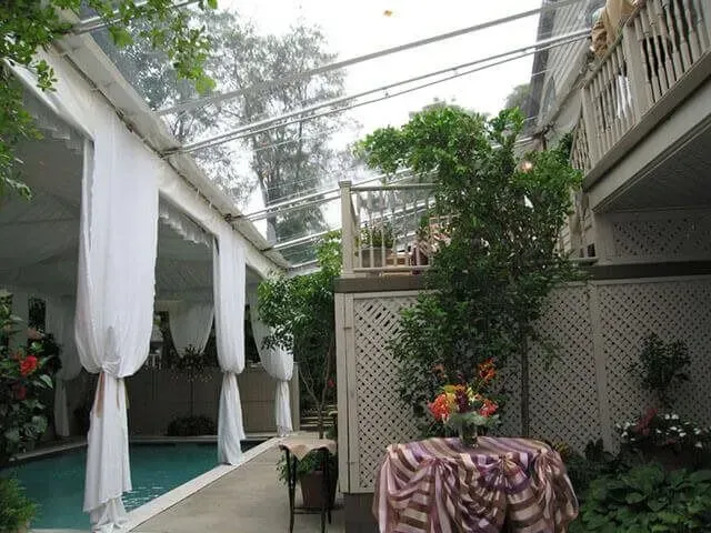 A pool deck enclosed by a glass roof and white curtains, featuring a round table with a striped cloth and potted plants.