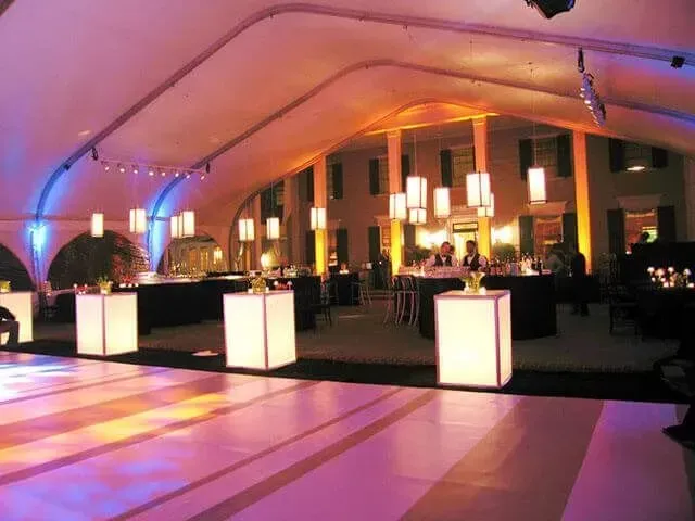 A lit-up dance floor in a tented event space featuring glowing square tables and warm pendant lighting at dusk.