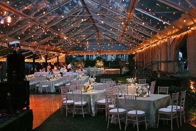 An outdoor evening wedding reception set up inside a clear-topped tent decorated with warm string lights.