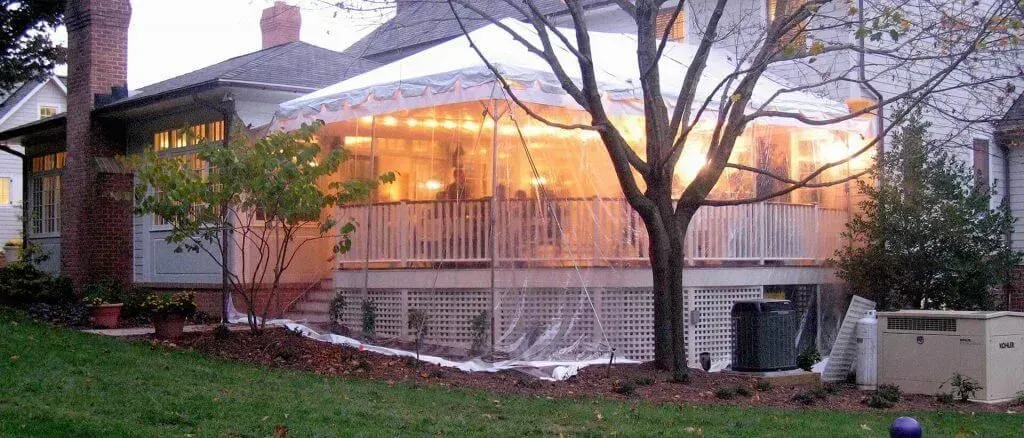 A residential deck enclosed in clear plastic sheeting, illuminated from within by warm lights, viewed from a lawn.