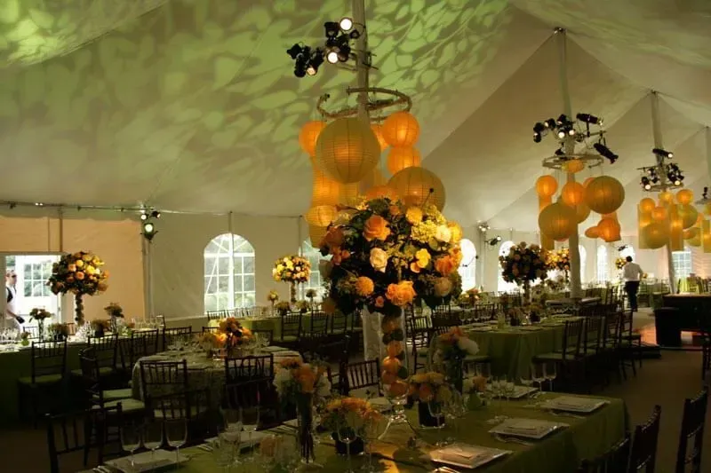 A formal event tent with round tables, yellow floral centerpieces, hanging paper lanterns, and green tablecloths.