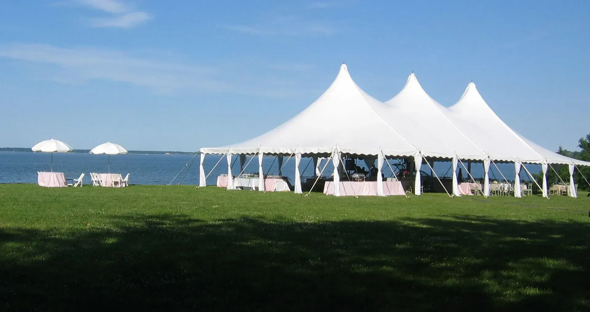 A large white event tent and umbrellas set up on a grassy lawn beside a body of water under a clear blue sky.