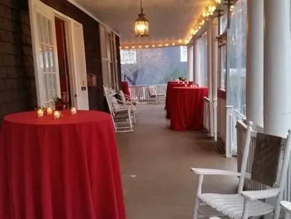 A long, covered porch featuring red-clothed round tables, white rocking chairs, and warm string lighting.