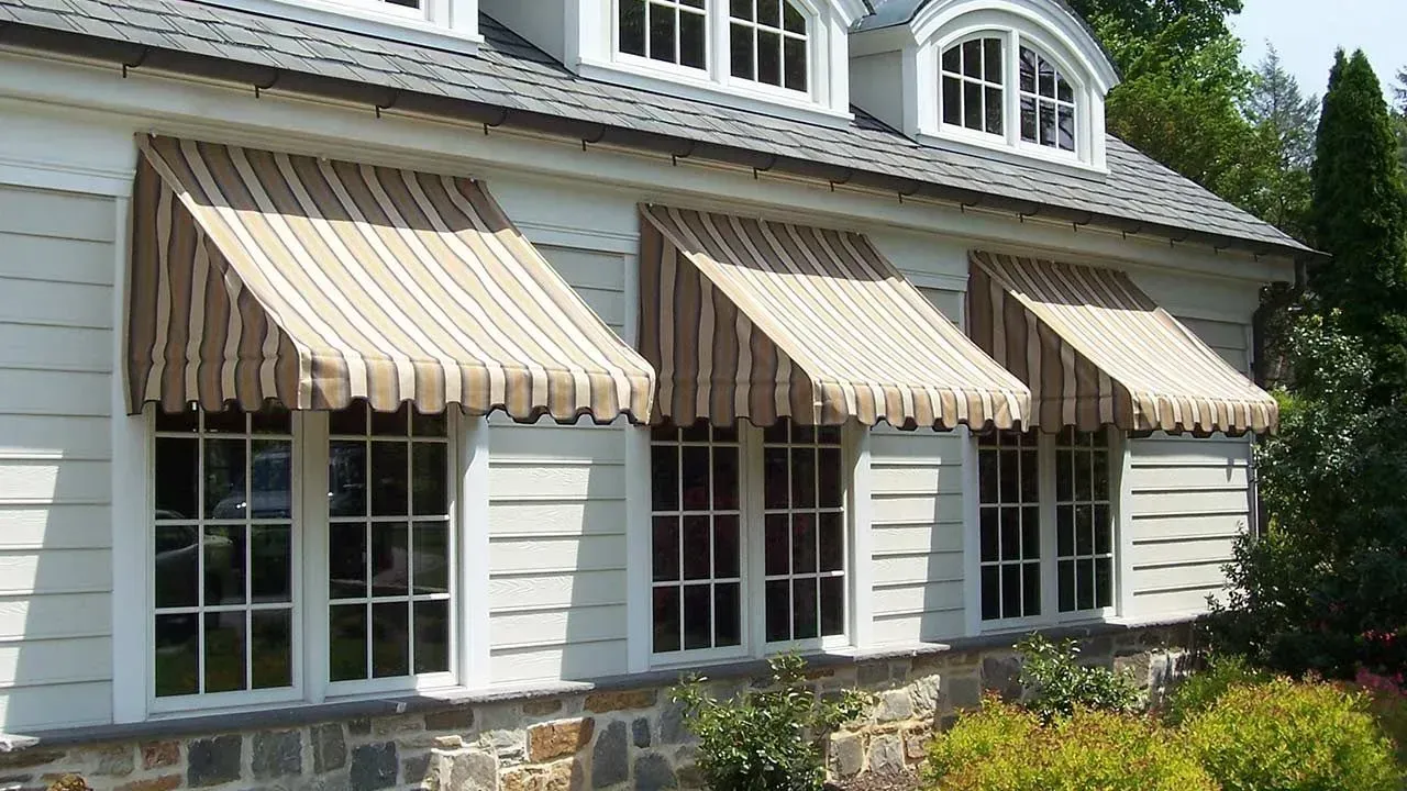 Three tan and white striped fabric awnings over the windows of a white-sided house with a stone foundation.