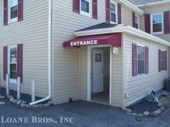Tan siding building with a maroon entrance awning and shutters, plus a white door and several tall bollards.