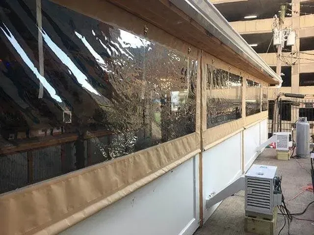 Outdoor structure with clear vinyl weather curtains and white lower panels, featuring an external heating unit nearby.