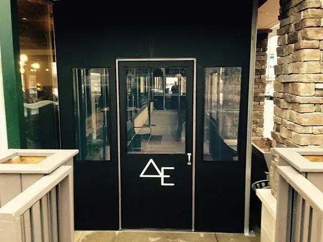 Entrance to a restaurant with a black door featuring a white triangle-and-E logo, flanked by two glass panels.