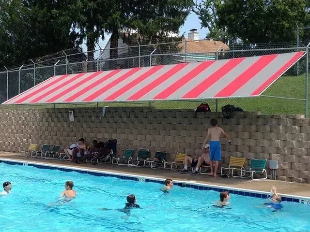 Children swim in an outdoor pool beneath a red and white striped shade canopy next to a retaining wall and lounge chairs.