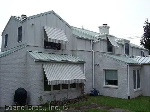A two-story gray brick house with green metal roofs and several striped window awnings on a grassy lawn.