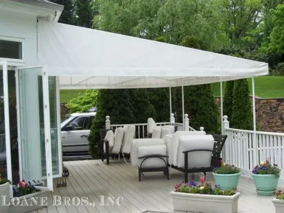 A white patio awning extends over a wooden deck with white chairs and potted flowers, viewed from a doorway.
