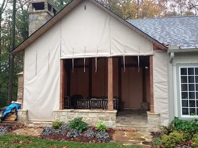 A covered stone patio features beige privacy curtains partially rolled up, revealing outdoor furniture and wooden posts.