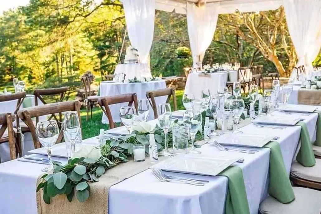 An outdoor wedding reception with long dining tables set with white tablecloths, greenery, and glassware under a tent.