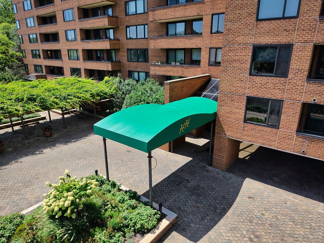 Brick apartment building entrance with green awning over a paved courtyard and trees nearby