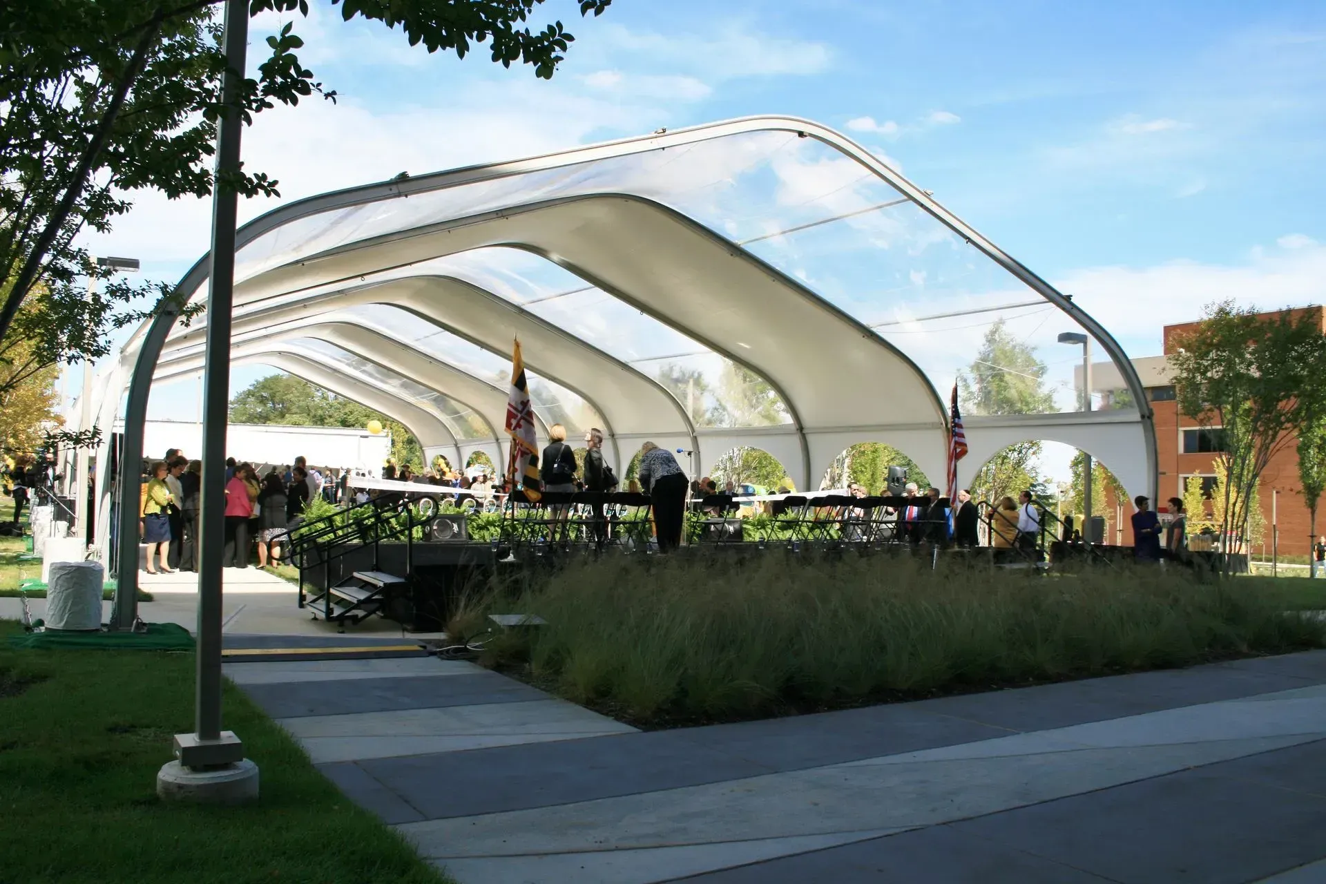 People gather under a large, arched white outdoor event tent on a sunny day with trees and a brick building nearby.