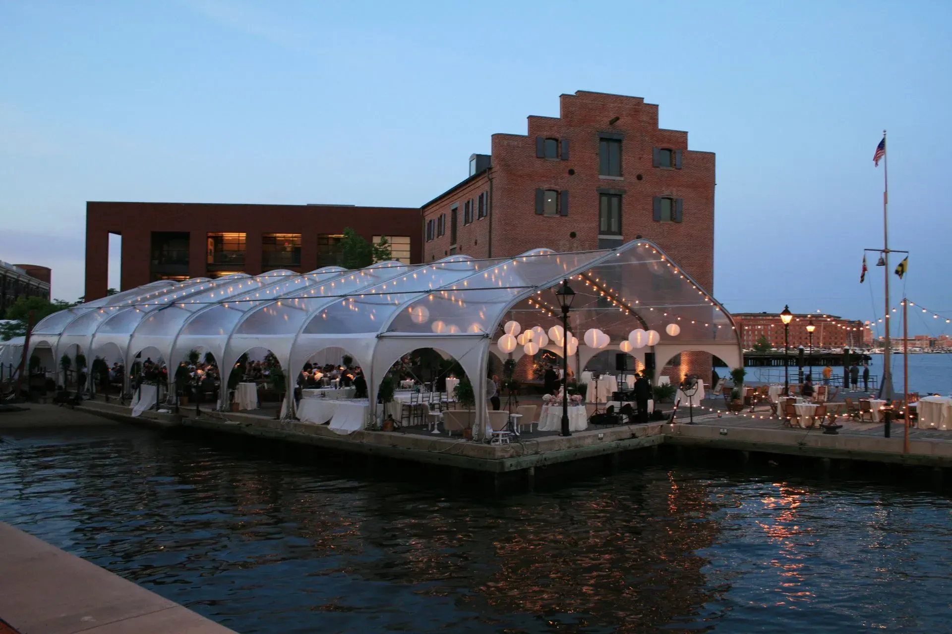 A clear, arched event tent on a waterfront pier at dusk, featuring interior lighting near a historic brick building.