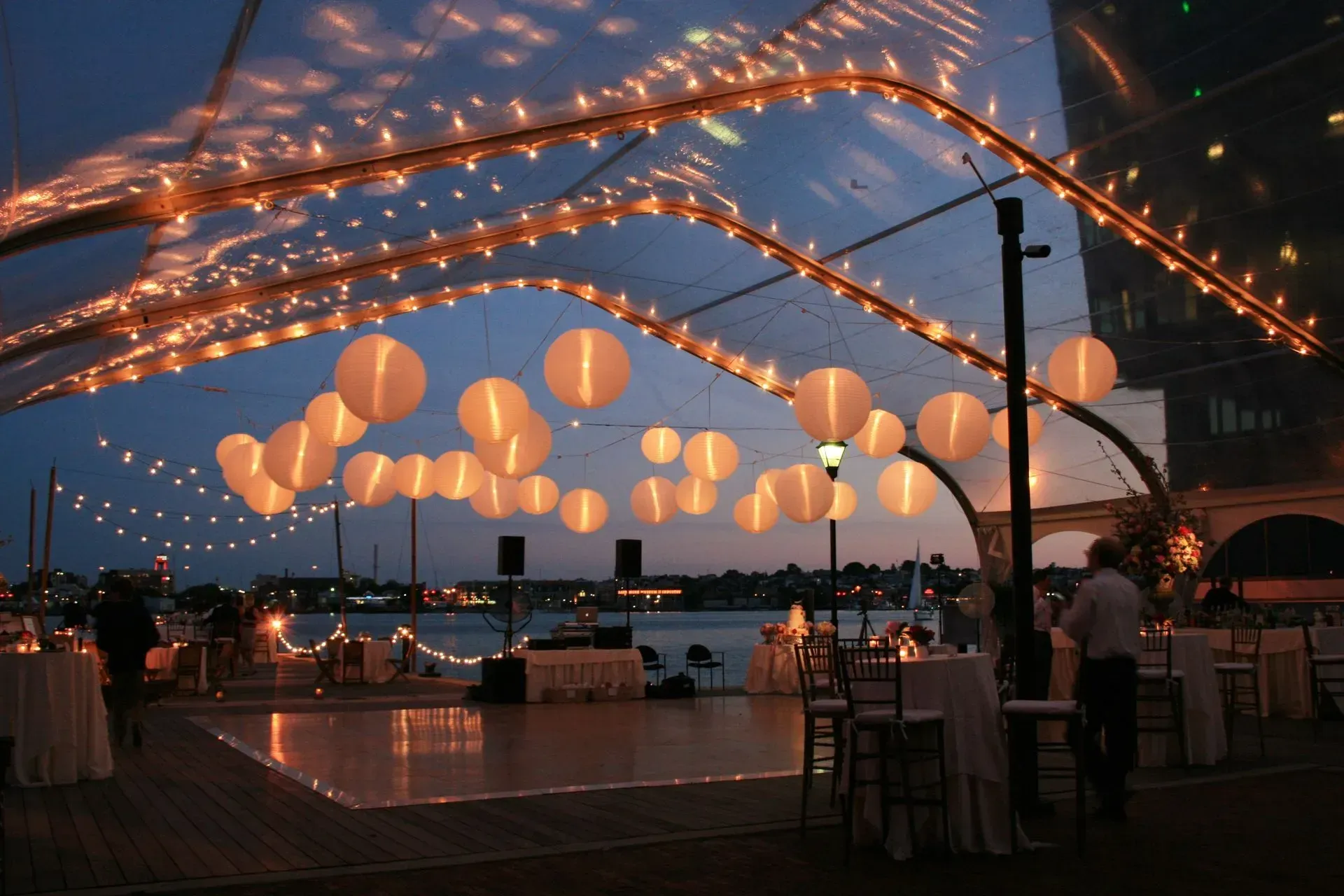 An outdoor evening event with a dance floor under a transparent tent canopy lit by warm string lights and paper lanterns.