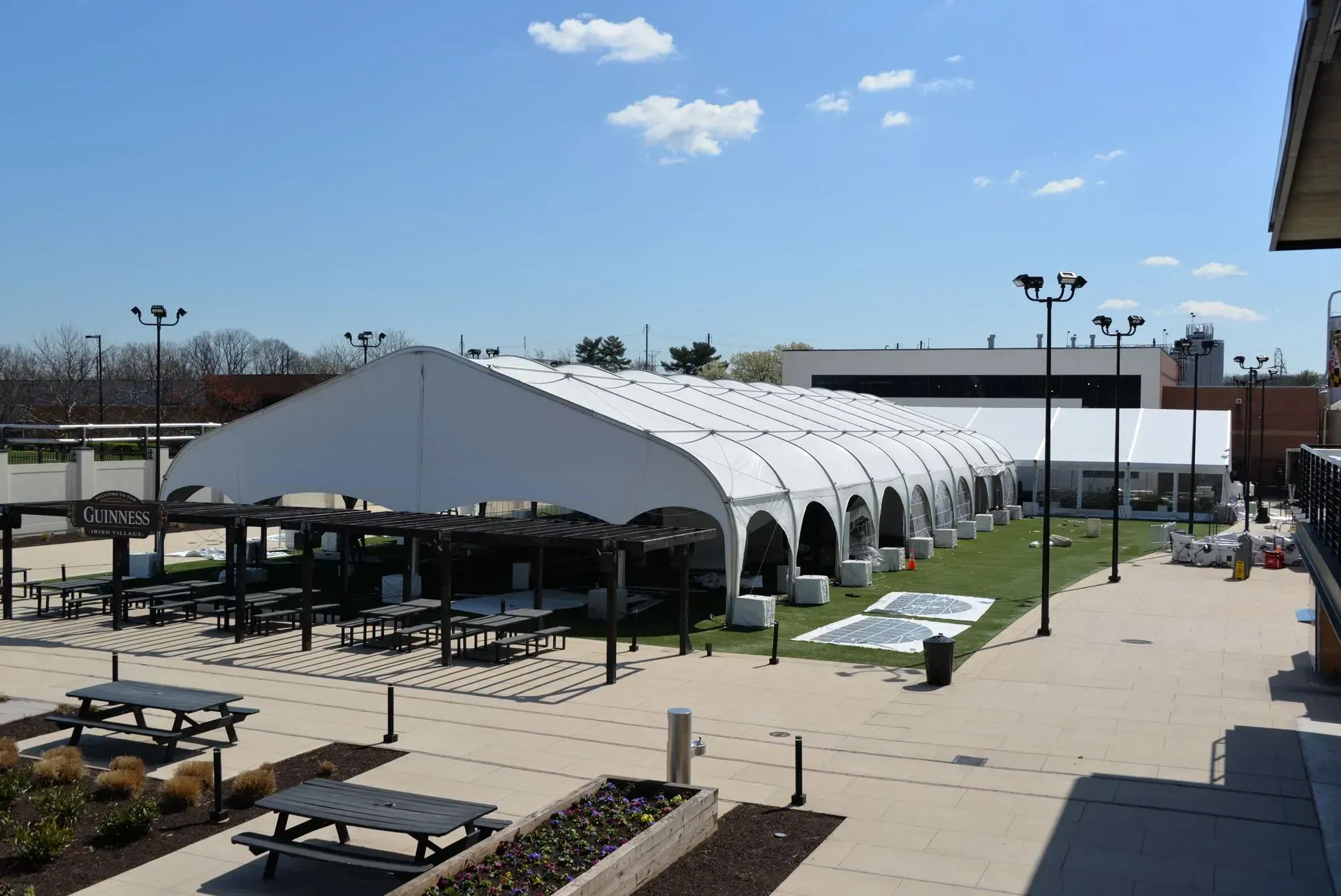 A large white event tent on a paved courtyard with outdoor picnic tables, bright lighting, and a clear blue sky.