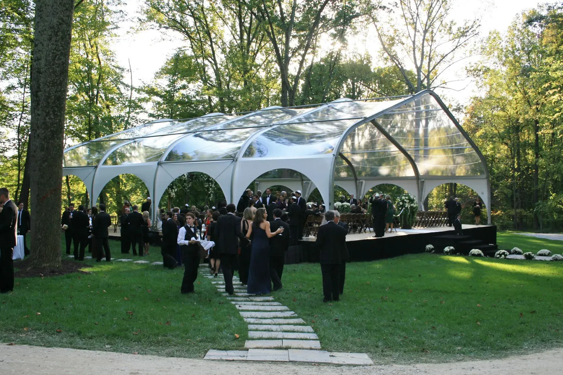 A group of people in formal wear gathers at an outdoor event under a large, arched, clear-roofed tent in a wooded area.
