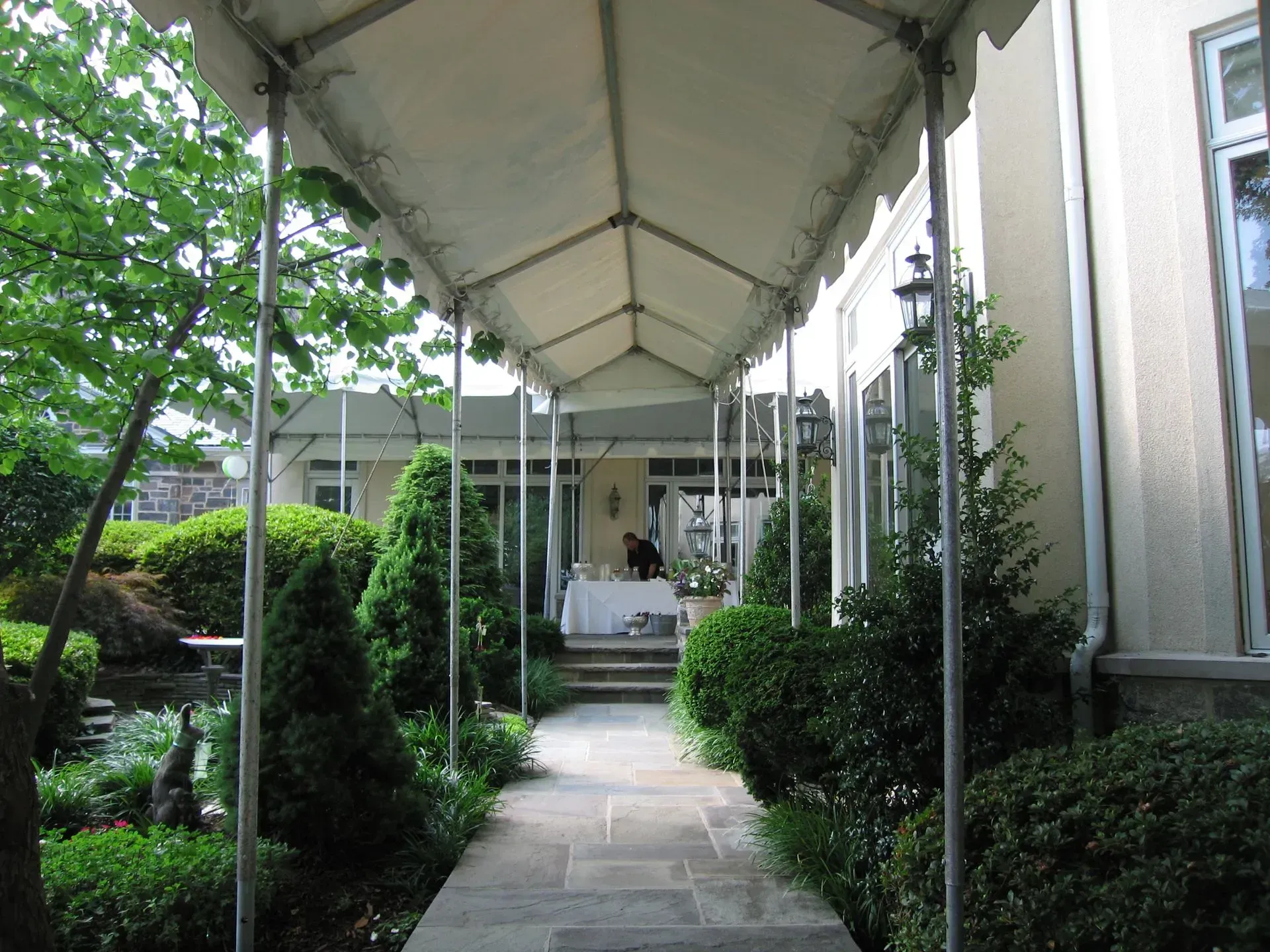A stone walkway covered by a tent canopy leads to an outdoor patio with lush green landscaping.