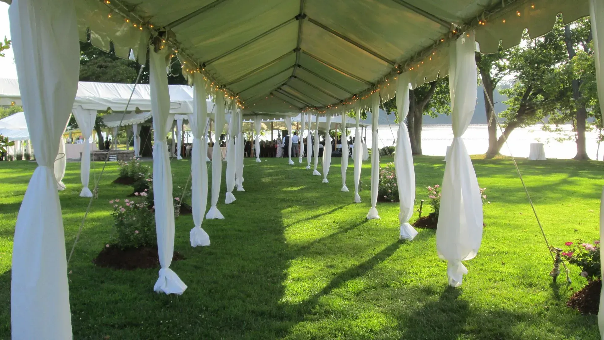 A long, white-canopied walkway draped with white fabric leads across a lush green lawn toward a sunlit background.