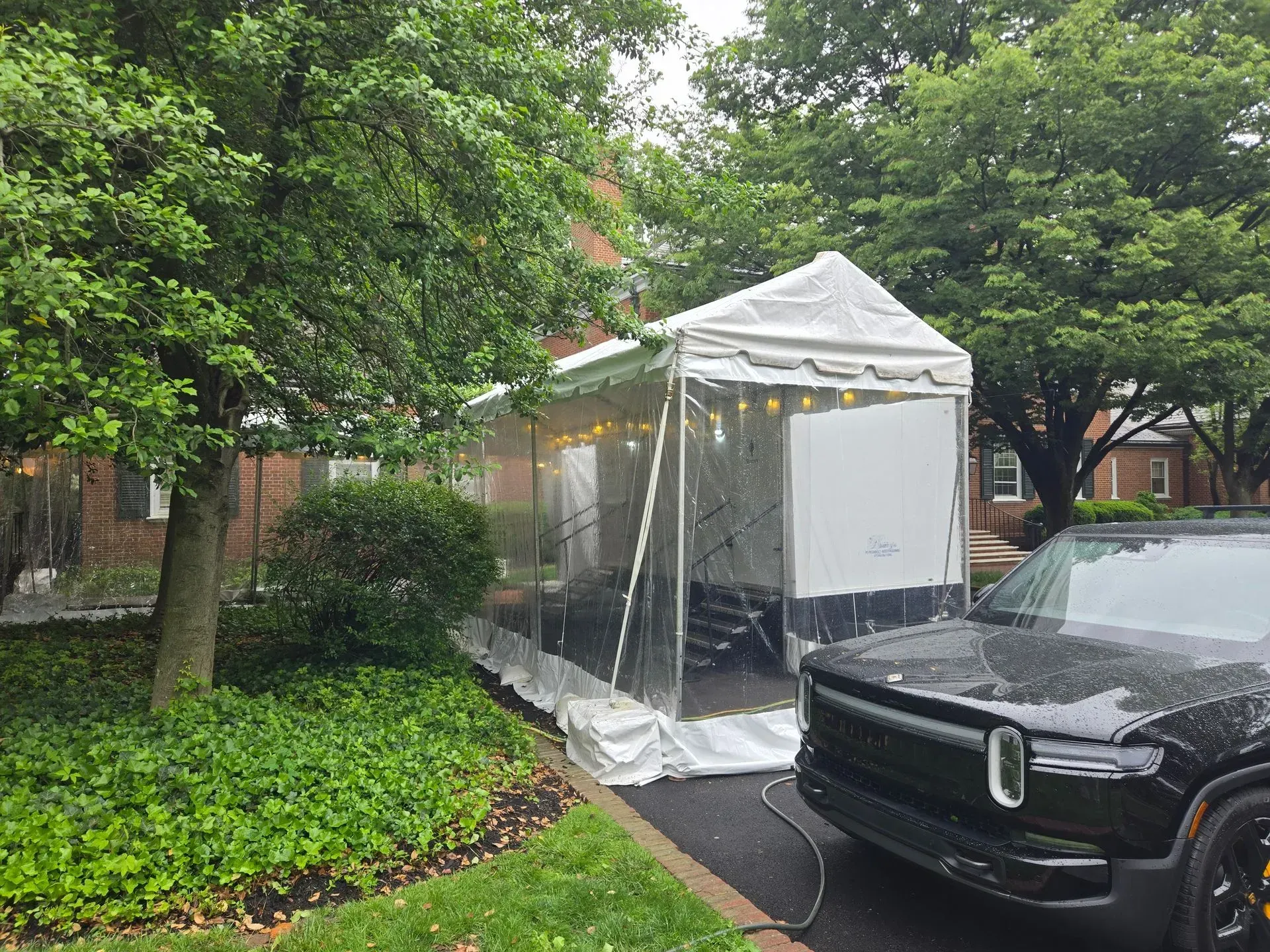 A white, peaked canopy tent stands on a driveway next to a black electric truck, near a grassy lawn and large trees.