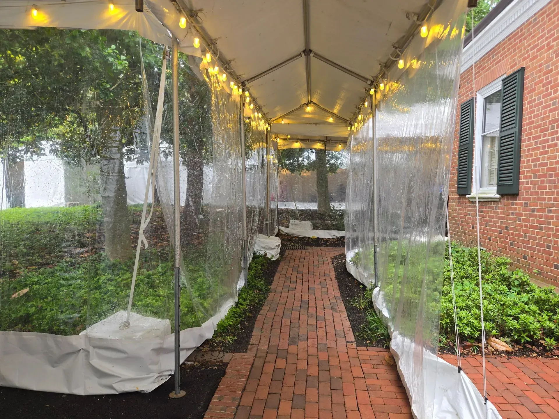 A covered walkway with clear plastic walls and string lights, leading over a red brick path next to a brick building.