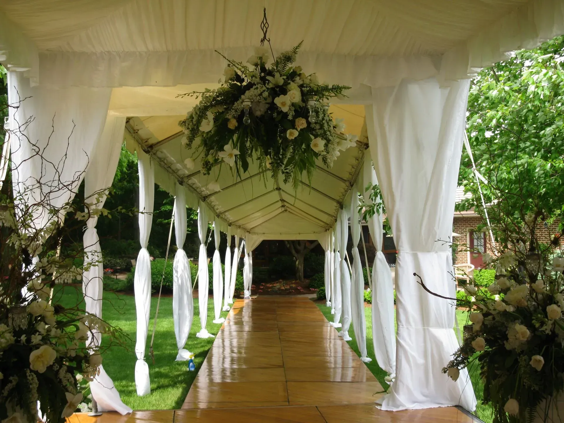 A wedding aisle featuring a white-draped canopy, floral arrangements, and a wooden pathway in a green outdoor garden.