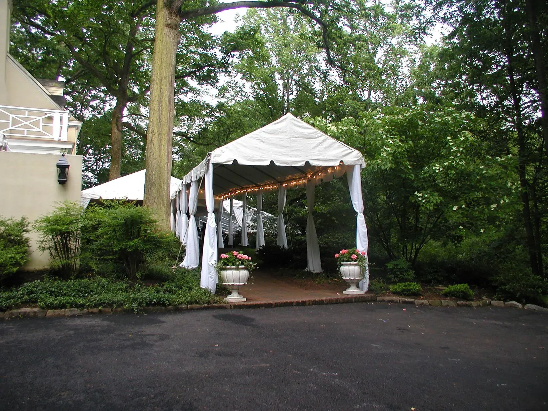 A white event tent stands on a paved driveway next to a building, framed by trees, with floral planters at the entrance.