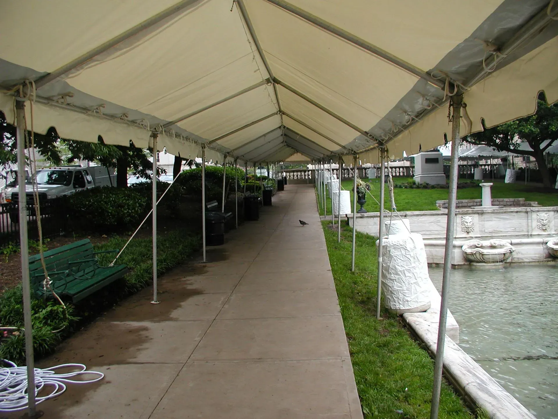 A covered walkway with a white canopy runs along a paved path, next to a park bench and a stone fountain.