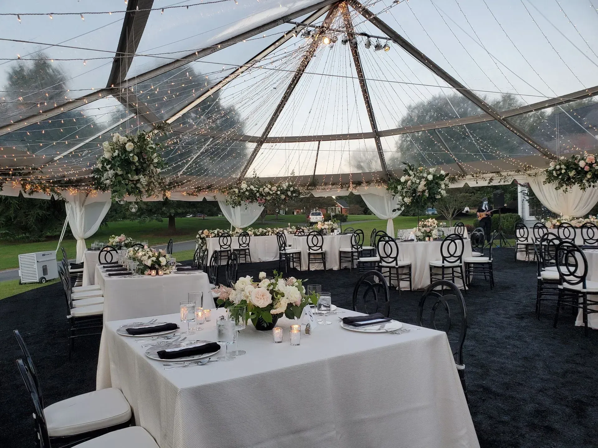 Tables set for a wedding reception under a clear-roofed tent with string lights, white linens, and dark carpeting.