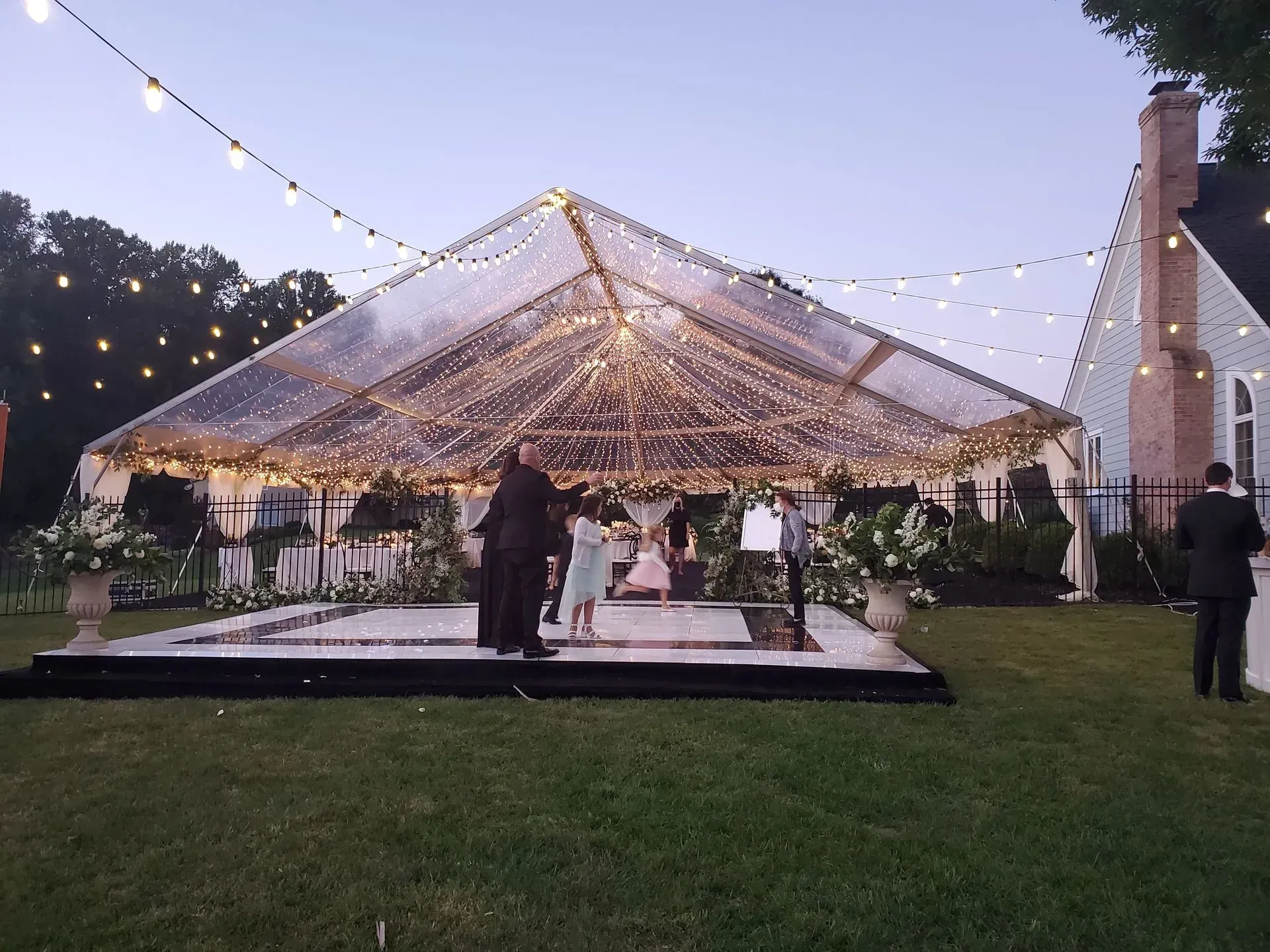 A clear, lit-up marquee tent at twilight with guests dancing on a platform outdoors next to a house.