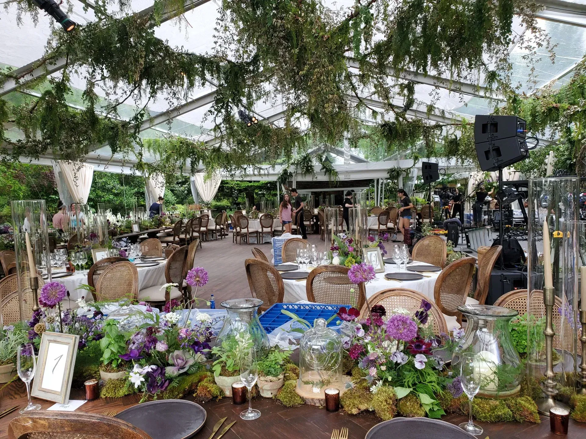 An outdoor event under a glass-roofed marquee with rustic tables, wicker chairs, and lush floral centerpieces.