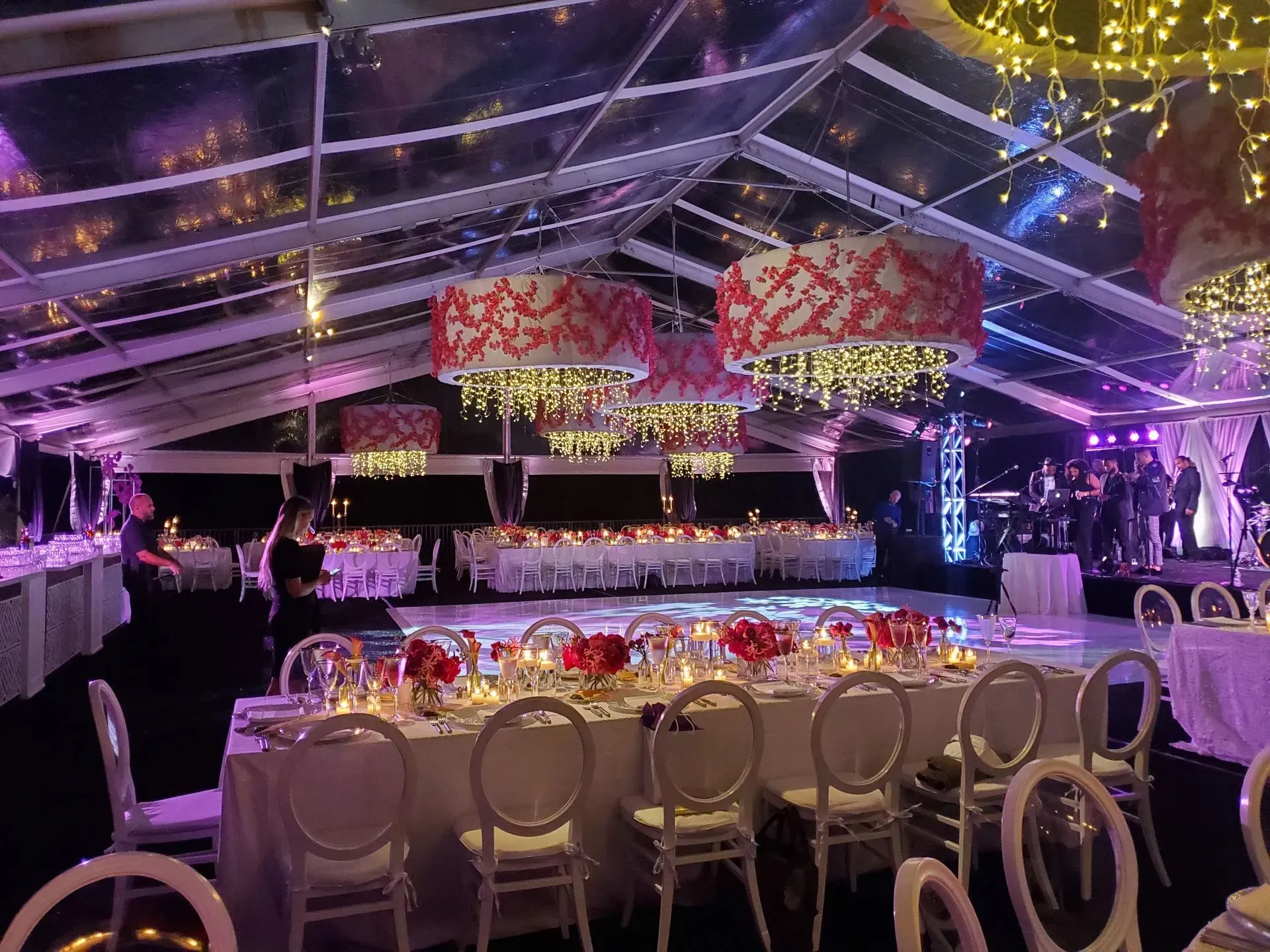 A banquet hall with long tables, white chairs, and large red-patterned drum chandeliers under a clear-roofed event tent.