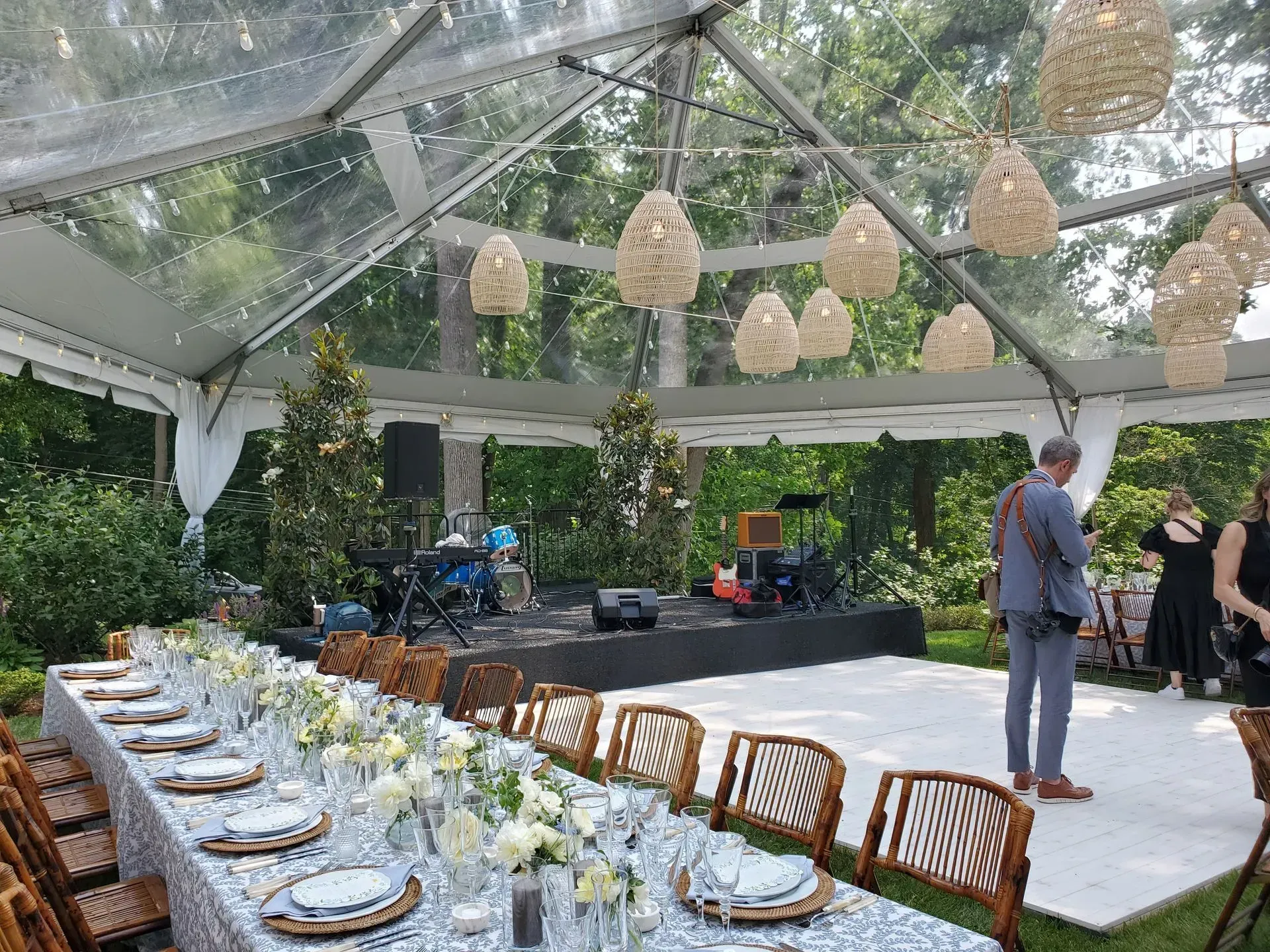 A clear tented reception with a long dining table, decorative hanging lamps, a band stage, and a white dance floor.