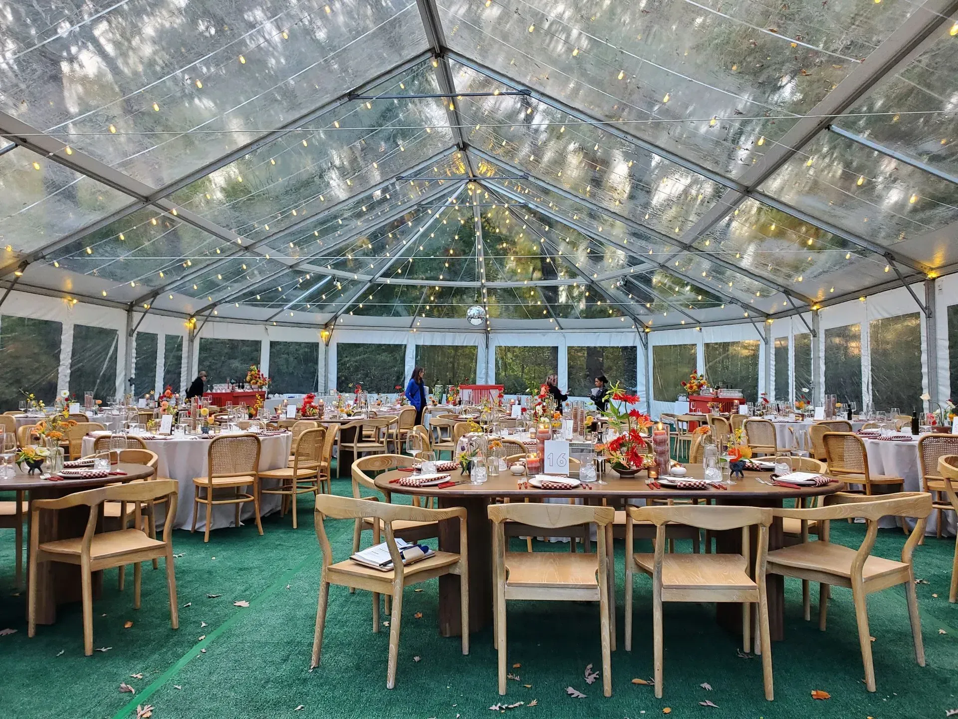 A clear-roofed event tent set for a dinner, featuring wooden chairs, round tables, and string lights over green flooring.