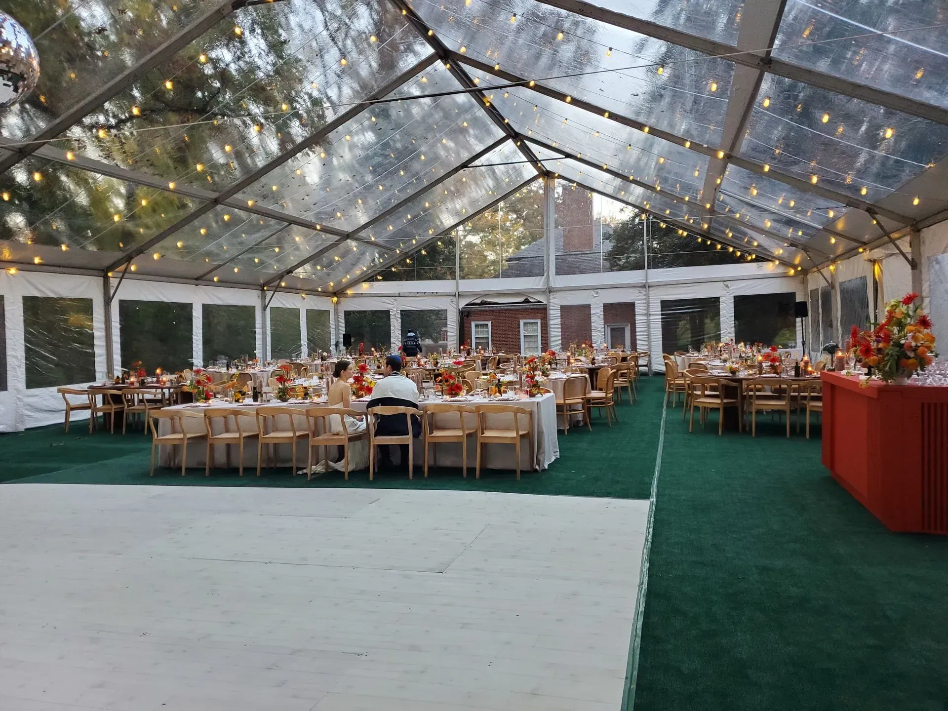 A clear-roofed event tent with dining tables, string lights, and a dance floor, set up on a green carpet.