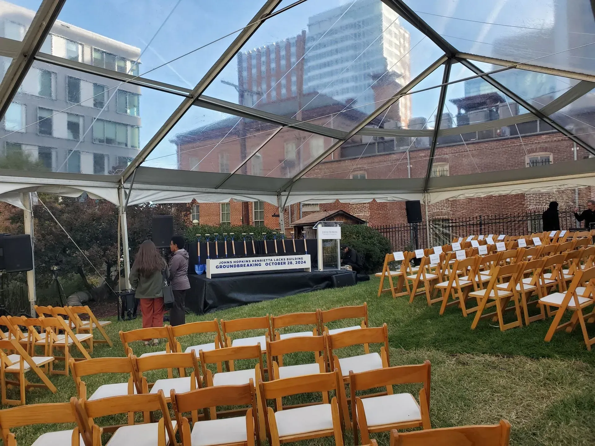 Rows of wooden folding chairs set up for an outdoor event under a clear-roofed tent, with city buildings in the background.